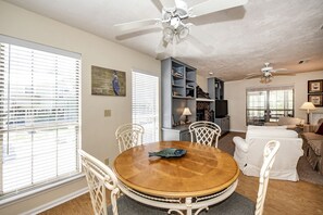A dining area with a round wooden table, white chairs, a ceiling fan, and a bookshelf.