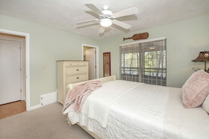 Une chambre à coucher comprenant un lit, une commode, un ventilateur de plafond, une fenêtre avec des stores et une porte.