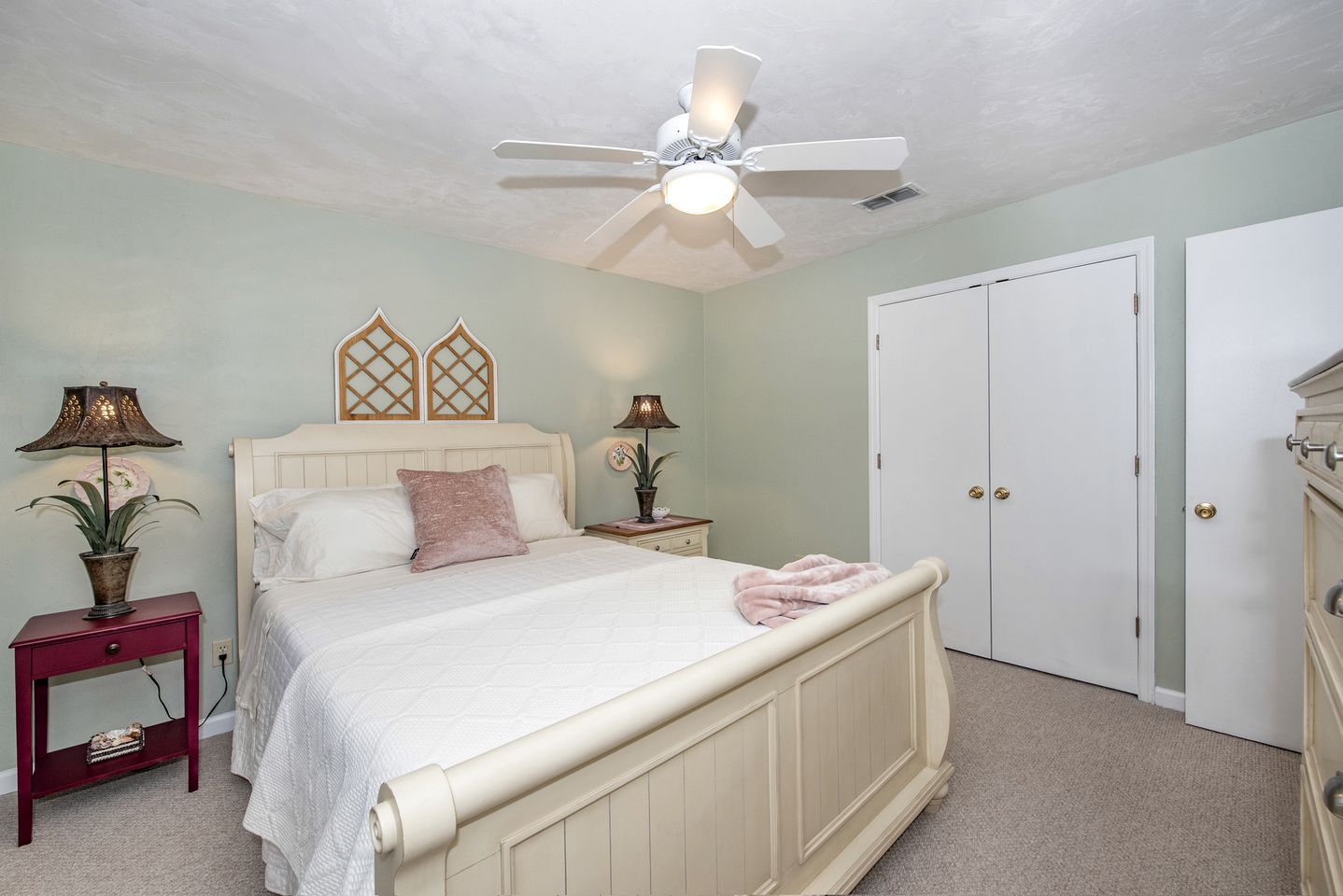 Une chambre à coucher comprenant un lit, deux tables de chevet, un ventilateur de plafond, une armoire et une porte.
