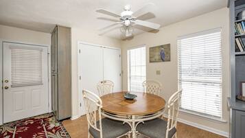 A dining room with a round wooden table, white chairs, a ceiling fan, and a bookshelf.