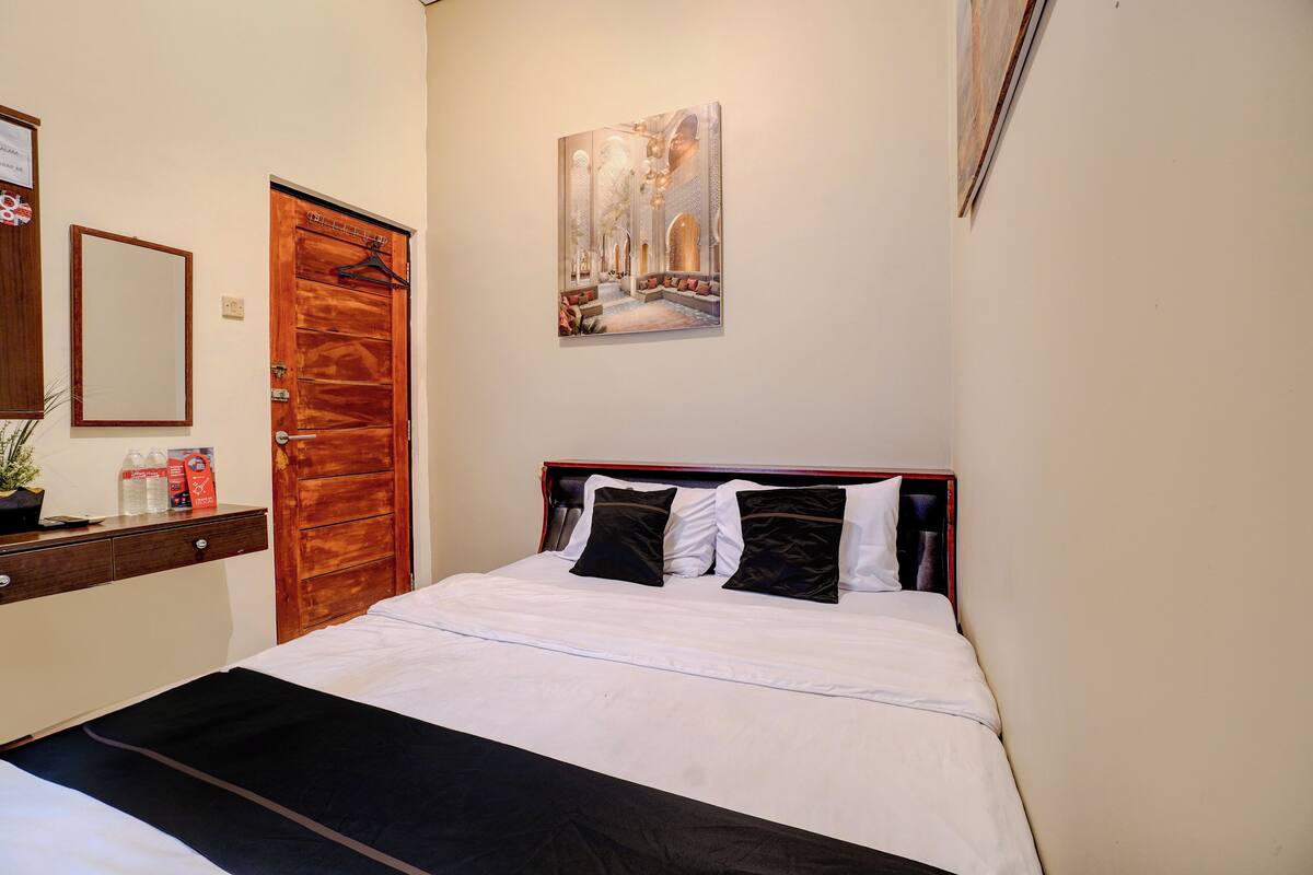 A hotel room with a bed, a wooden door, a wall-mounted painting, and a small desk with toiletries.