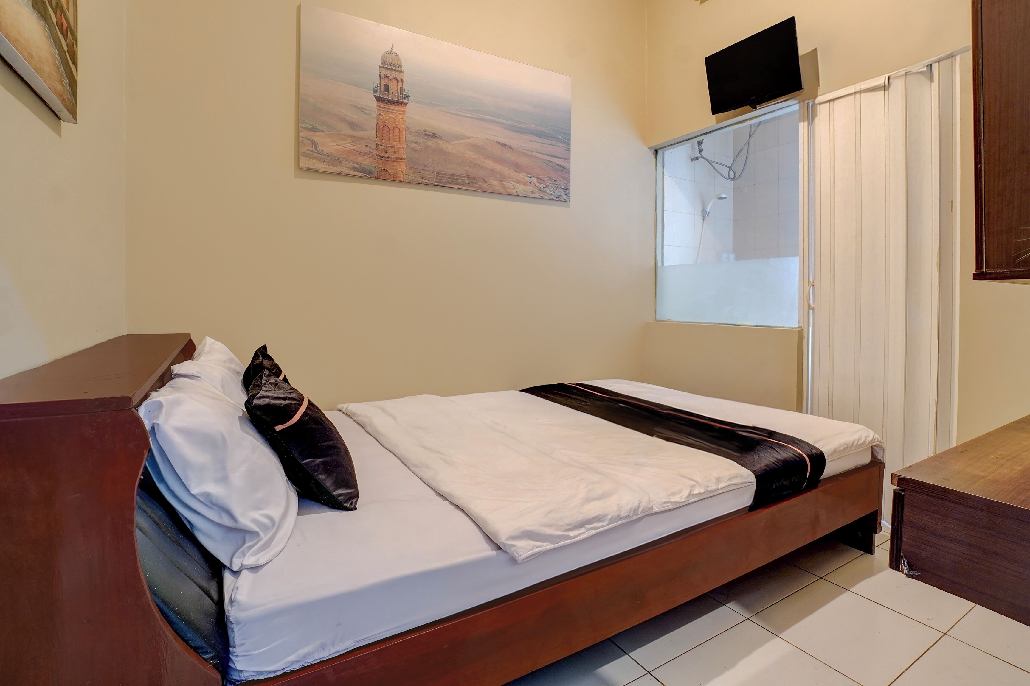 A single bed with white bedding and a black accent, a wooden headboard, a painting of a tower on the wall, a TV mounted above the bed, and a window with blinds.