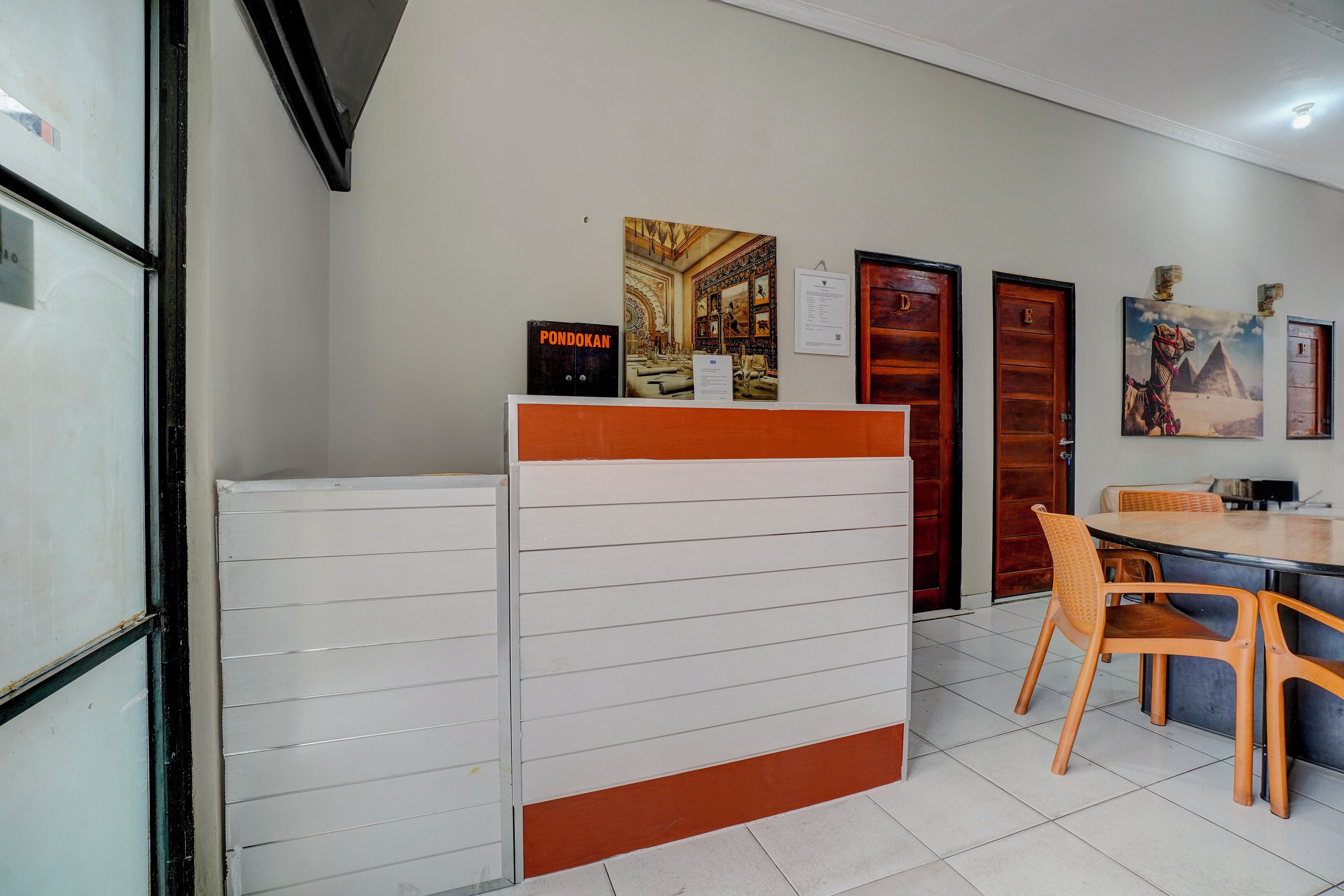 Une salle d’attente avec un comptoir blanc, des chaises en bois et une table. Des tableaux encadrés ornent le mur, et une porte est marquée de la lettre « D ».