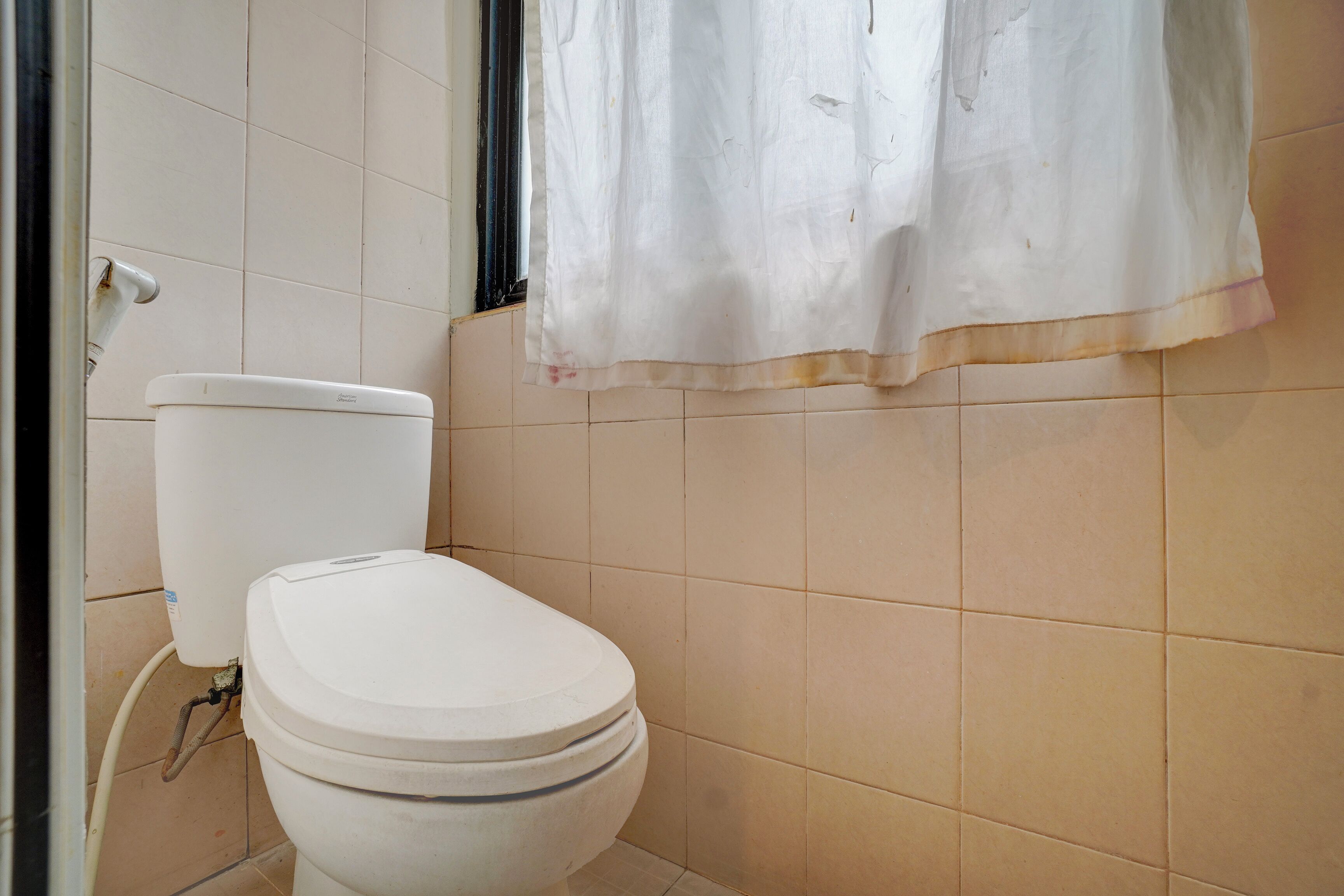 A bathroom with a toilet, a visible pipe, and a window with a curtain.