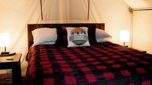 A bed with a red and black checkered bedspread, two white pillows, and a pillow with a mountain and forest design. There is a bedside table with a lamp and a wooden nightstand.