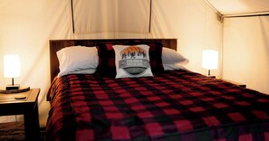 A bed with a red and black checkered bedspread, two white pillows, and a pillow with a mountain and forest design. There is a bedside table with a lamp and a wooden nightstand.