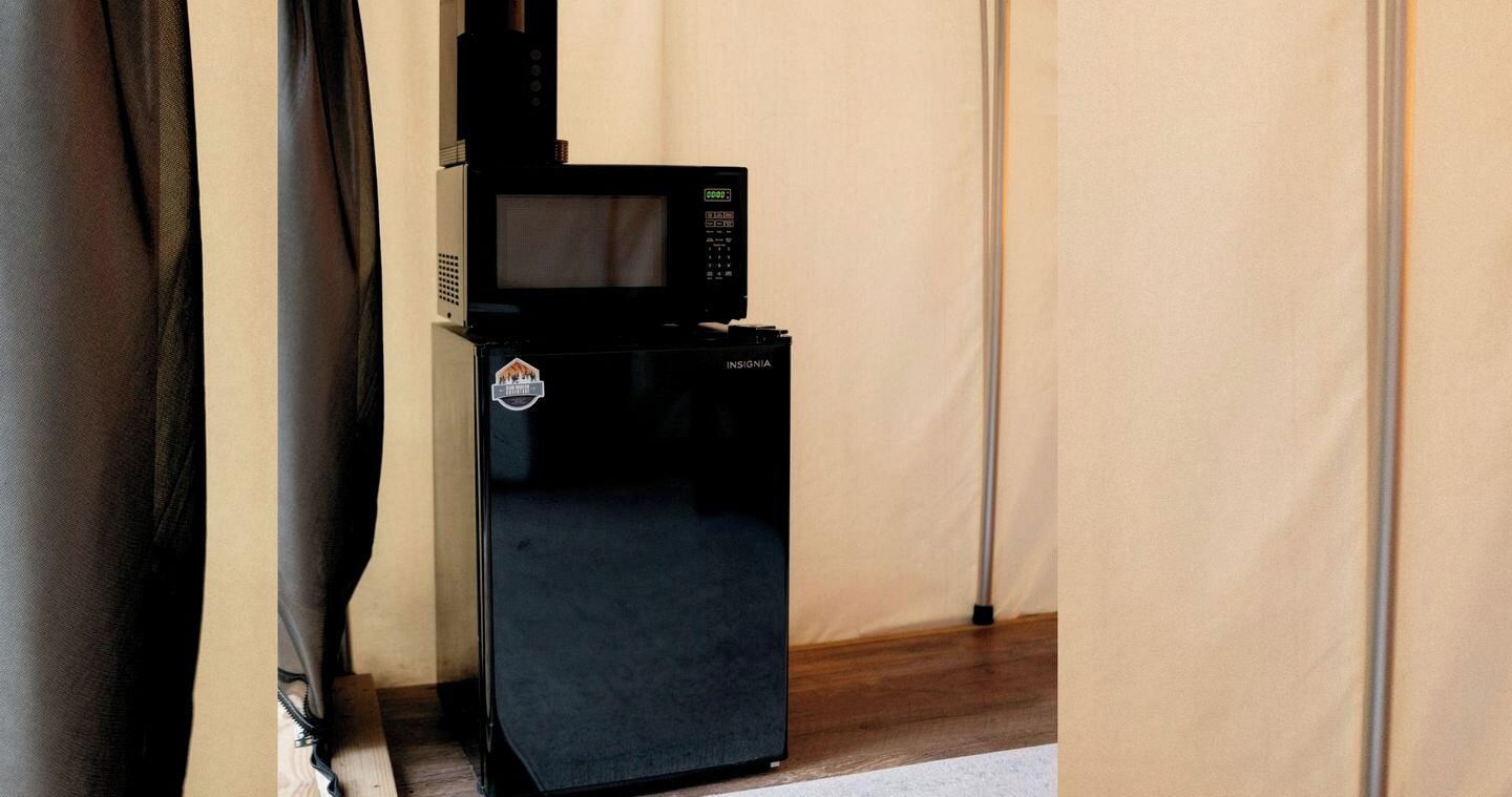 A black refrigerator with a microwave on top, placed in a corner of a room with beige walls and a wooden floor.