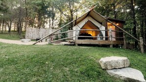 A rustic wooden cabin with a pitched roof, surrounded by a wooden fence, set in a forested area with a gravel path leading to it.