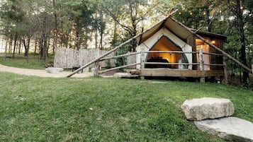 A rustic wooden cabin with a pitched roof, surrounded by a wooden fence, set in a forested area with a gravel path leading to it.