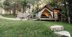 A rustic wooden cabin with a pitched roof, surrounded by a wooden fence, set in a forested area with a gravel path leading to it.