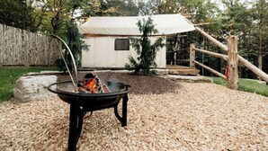 A campfire in a black metal fire pit surrounded by gravel, with a tent and wooden fence in the background.