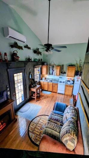A spacious living room with a fireplace, a sofa, a dining table, and a kitchen area with wooden cabinets and a stove. - Sparta Serenity: Charming Cabin with Hot Tub and Scenic Farm Views in TN (Sparta)