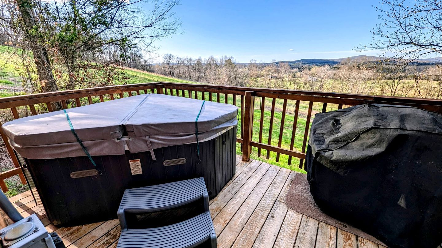 A wooden deck with a hot tub, a covered seating area, and a large black object, likely a storage or seating unit, overlooking a rural landscape.