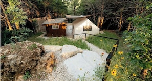 Una cabaña rústica con un cobertizo pequeño, rodeada de un paisaje natural con rocas y flores silvestres.
