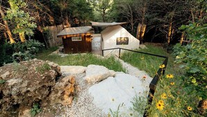 Eine ländliche Outdoor-Szenerie mit einer kleinen Holzhütte und einem Zelt, umgeben von natürlicher Vegetation und Felsen.