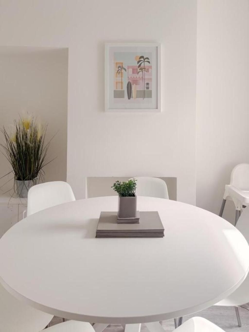 Een ronde tafel met een bloempot en een stapel boeken in het midden, omringd door witte stoelen.