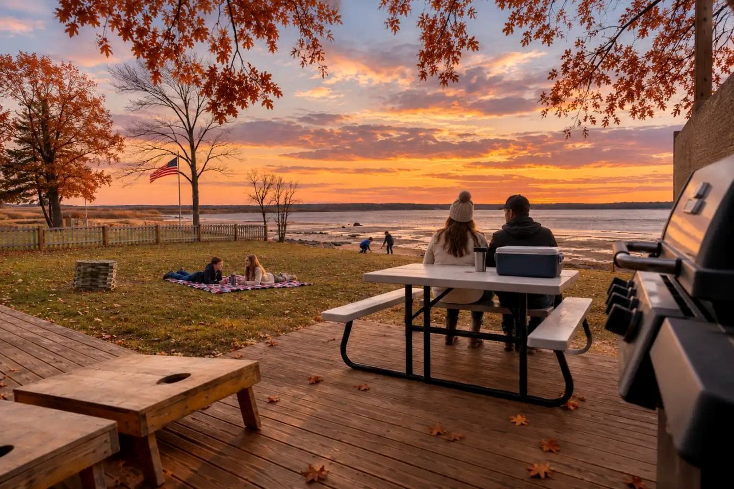 A deck with picnic tables and a grill, overlooking a beach at sunset.