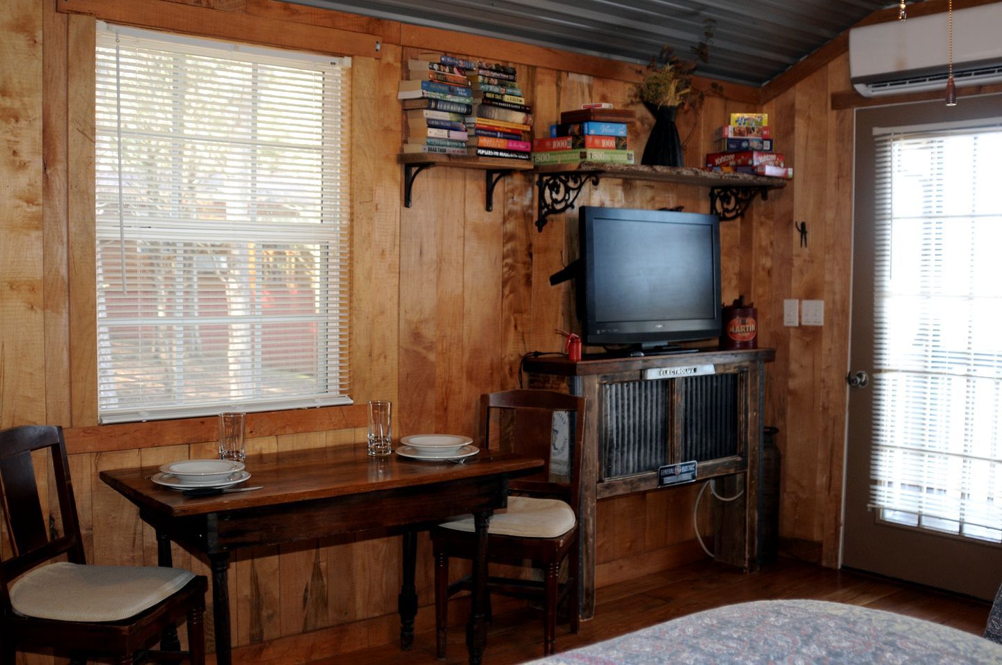 A wooden-paneled room with a television, a dining table with chairs, and a window with blinds.