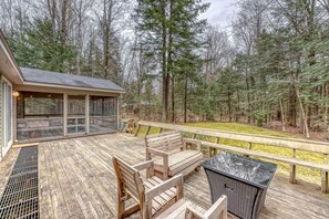 A wooden deck with outdoor seating, a covered area with windows, and a forested backdrop.