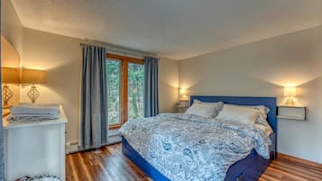 A bedroom with a bed, bedside lamps, a window with curtains, a nightstand, and a wooden floor.