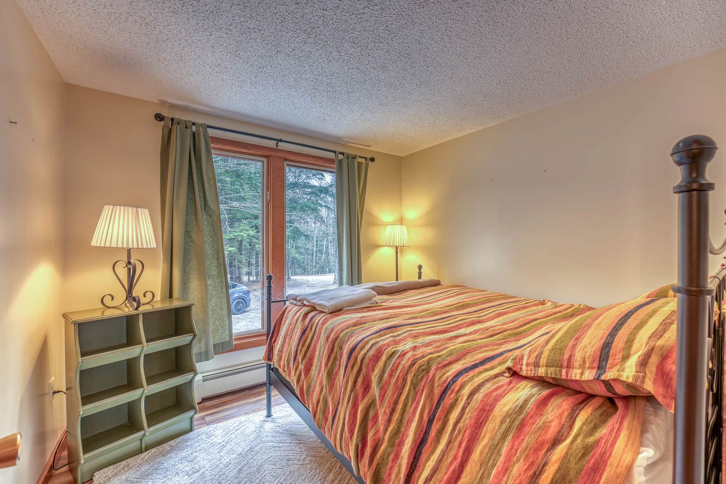 Un dormitorio con cama, mesita de noche, lámpara, ventana con cortinas y vistas al exterior.