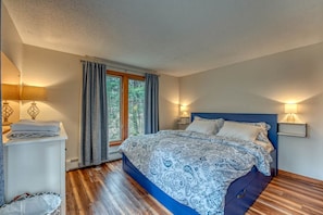 A bedroom with a bed, bedside lamps, a window with curtains, a nightstand, and a wooden floor.