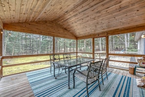A covered wooden deck with a glass table and chairs, surrounded by a forest view.