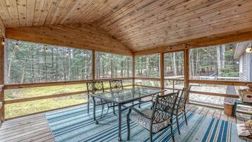 A covered wooden deck with a glass table and chairs, surrounded by a forest view.