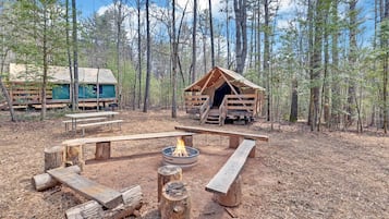 A rustic campsite with a fire pit, picnic tables, and a wooden cabin.