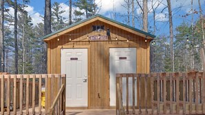 Una cabaña de madera con un letrero que dice 'BATHHOUSE' sobre la entrada, dos puertas identificadas como 'Baño de hombres' y 'Baño de mujeres', y una cerca de madera que rodea el área.