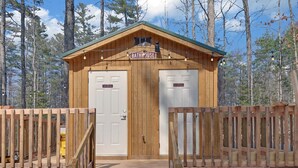 Eine Holzhütte mit einem Schild, auf dem 'BADEHAUS' steht, zwei Türen mit den Aufschriften 'Herren-WC' und 'Damen-WC' sowie ein Holzzaun, der das Grundstück umgibt.
