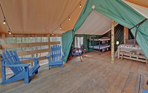Un rincón exterior acogedor con suelo de madera, una silla Adirondack azul, una mesita y una zona de dormitorio cubierta con una cama y una litera.