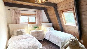 Ein Schlafzimmer mit zwei Betten, einem Nachttisch, einer Lampe, einem Teddybären und einem Fenster mit Vorhängen.
