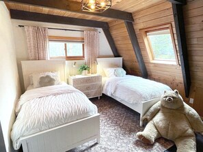 Ein Schlafzimmer mit zwei Betten, einem Nachttisch, einer Lampe, einem Teddybären und einem Fenster mit Vorhängen.