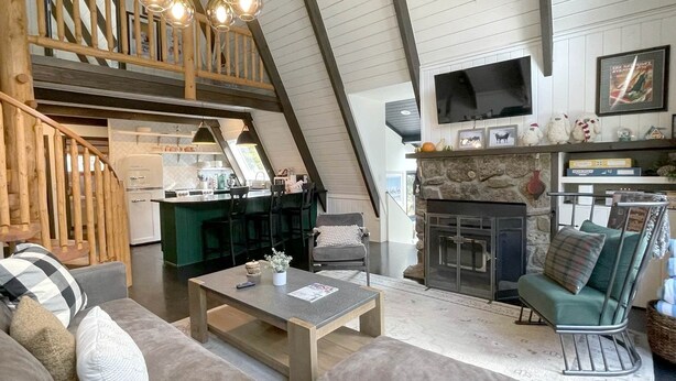 Ein Wohnbereich im Loft-Stil mit hohen Decken, freiliegenden Holzbalken, einem Steinkamin, einem Fernseher und einer Küche im Hintergrund.