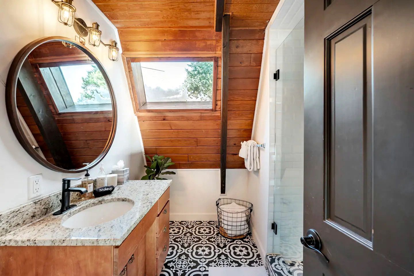 Un baño con techo de madera, un espejo grande, un lavabo con encimera de granito, una ventana pequeña y baldosas de suelo con diseño.