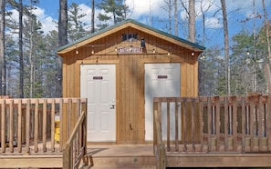 A wooden cabin with a sign reading 'BATHHOUSE' above the entrance, two doors labeled 'Men's Bathroom' and 'Women's Bathroom,' and a wooden fence surrounding the area.