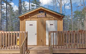Una cabaña de madera con un letrero que dice 'BATHHOUSE' sobre la entrada, dos puertas identificadas como 'Baño de hombres' y 'Baño de mujeres', y una cerca de madera que rodea el área.