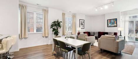 Un salon moderne avec un coin repas, comprenant une table blanche, des chaises vertes, un canapé et une grande fenêtre avec des rideaux.