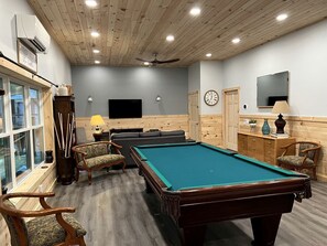 Une table de billard dans une pièce aux murs en bois, avec un ventilateur au plafond et un téléviseur.