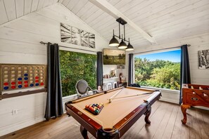 Une table de billard avec une queue de billard, une table de snooker et une table de baby-foot dans une pièce au plancher de bois et aux murs blancs.