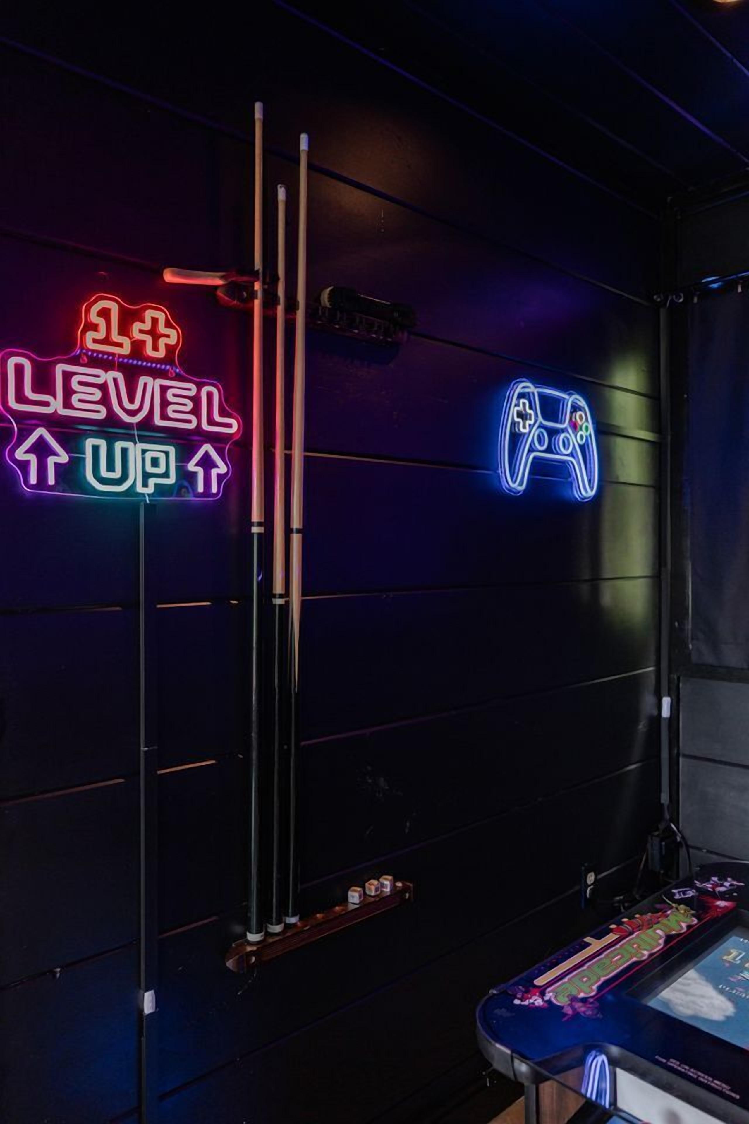 Ein Neon-Schild mit der Aufschrift 'LEVEL UP' und einem Symbol für einen Spielecontroller, ein vertikaler Pfosten und ein Tisch mit einem bunten Gegenstand darauf.