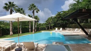 Ein Swimmingpool mit Liegestühlen und Sonnenschirmen, umgeben von Palmen und viel Grün.