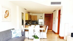 Ein modernes Wohnzimmer mit grauem Sofa, weißem Esstisch und Stühlen. Im Hintergrund ist eine Küche mit Holzschränken und einer Mikrowelle zu sehen.