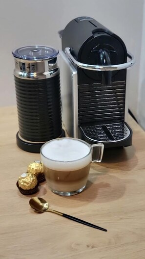 Een koffieapparaat, een thermoskan en een kop koffie met twee chocolades en een lepel op een houten tafel.