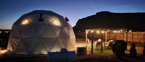 A geodesic dome tent with a circular skylight, a covered outdoor seating area with string lights, and a grill.