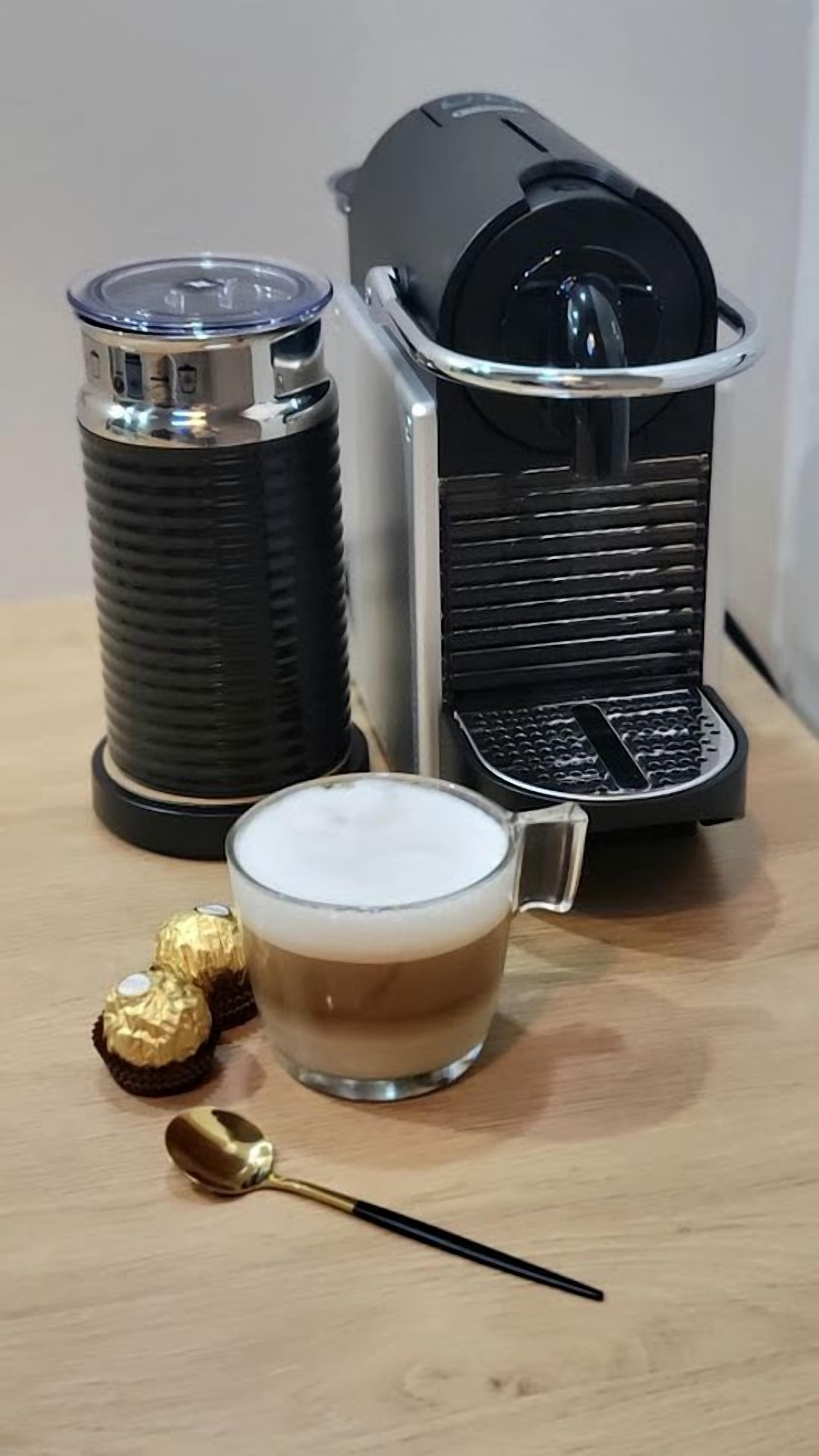 A coffee machine, a thermos, and a cup of coffee with two chocolates and a spoon on a wooden table.