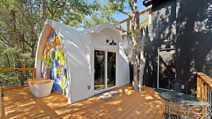 A wooden deck with a glass door entrance, a white wall with a colorful mural, and a metal table and chair.