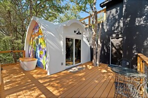 A wooden deck with a glass door entrance, a white wall with a colorful mural, and a metal table and chair.
