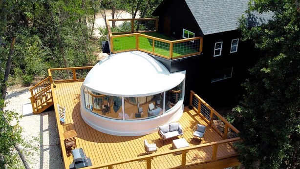 A circular building with a glass dome, a deck with seating, and a black house nearby.